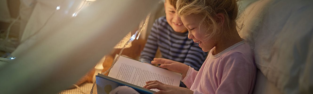 Two children reading a book under a blanket tent.